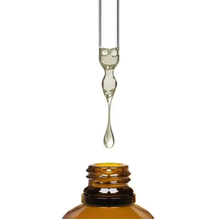 Glass dropper releasing a drop of clear oil above an open amber bottle